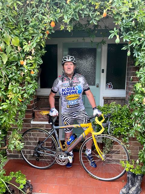 Ray wearing cycling gear and with his yellow mountain bike, stood in a doorway edged with a hedge. 